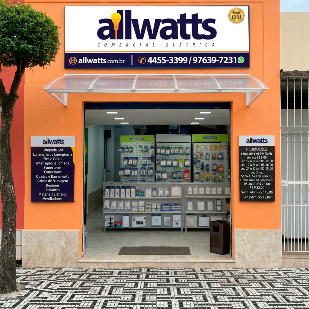 Visão frontal da fachada da loja Allwatts Comercial Elétrica, com parede alaranjada e entrada centralizada. Na parte superior, destaca-se a placa com o logotipo da Allwatts, o selo "Desde 1991", site e telefones de contato. Um toldo transparente protege a entrada. Nas laterais da porta, há dois quadros informativos da Allwatts com categorias de produtos e lista de promoções. O interior exibe balcões e expositores organizados, e o piso externo é de calçada em mosaico.