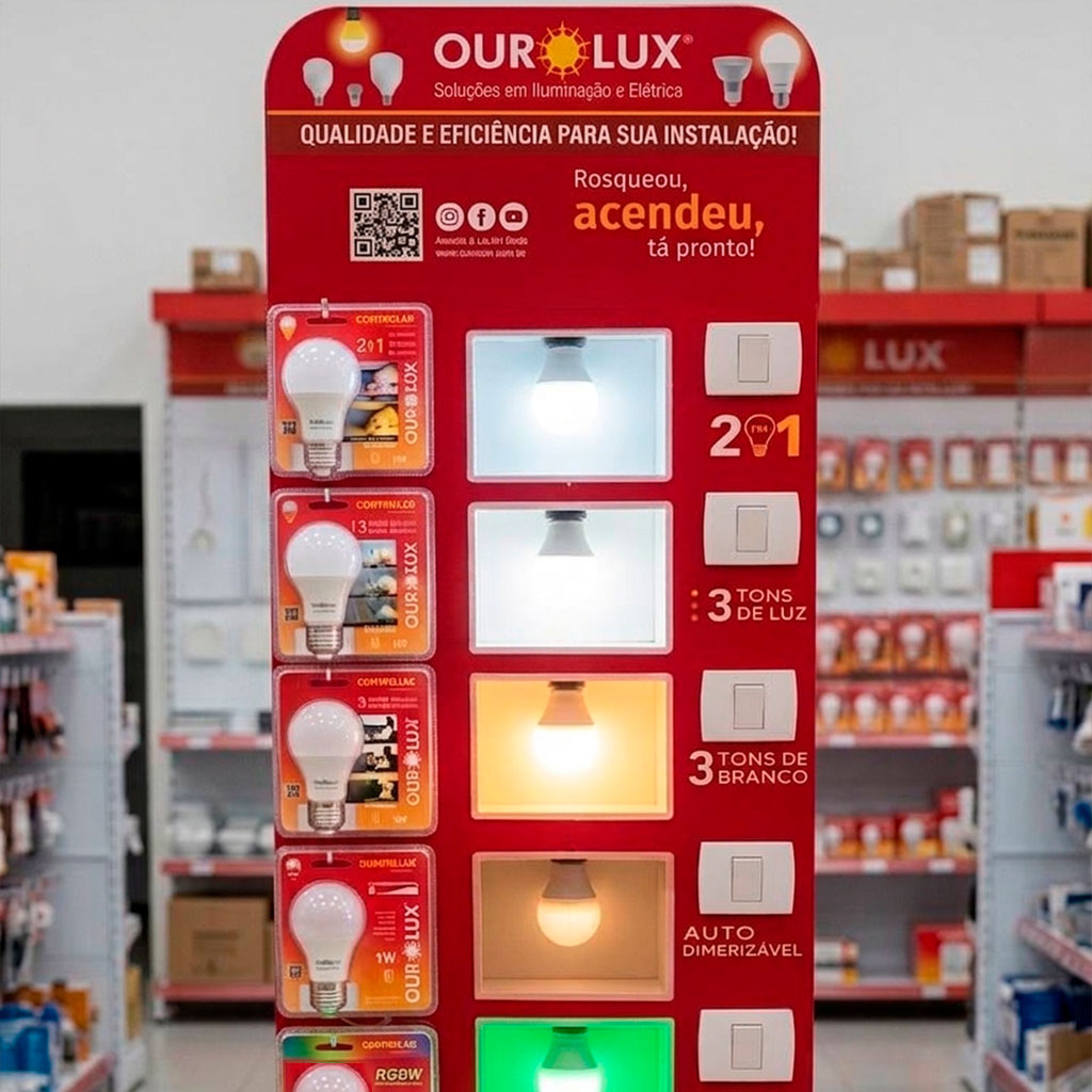 Visão frontal e detalhada de um display expositor vertical vermelho da marca Ourolux. No topo, o painel vermelho exibe a marca "OUROLUX - Soluções em Iluminação e Elétrica", a frase "QUALIDADE E EFICIÊNCIA PARA SUA INSTALAÇÃO!" e o slogan "Rosqueou, acendeu, tá pronto!" com QR code e redes sociais. O painel possui amostras de lâmpadas LED em embalagens no lado esquerdo, nichos iluminados ao centro mostrando diferentes temperaturas de cor e funções, e especificações no lado direito.